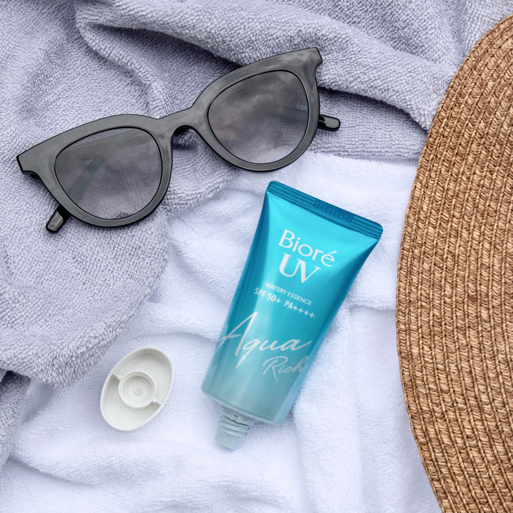 Biore UV Aqua Rich Watery Essence SPF50+/PA++++ tube beside sunglasses on a soft towel background.