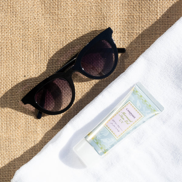 Canmake Mermaid Skin Gel UV sunscreen tube beside stylish black sunglasses on a beach towel.